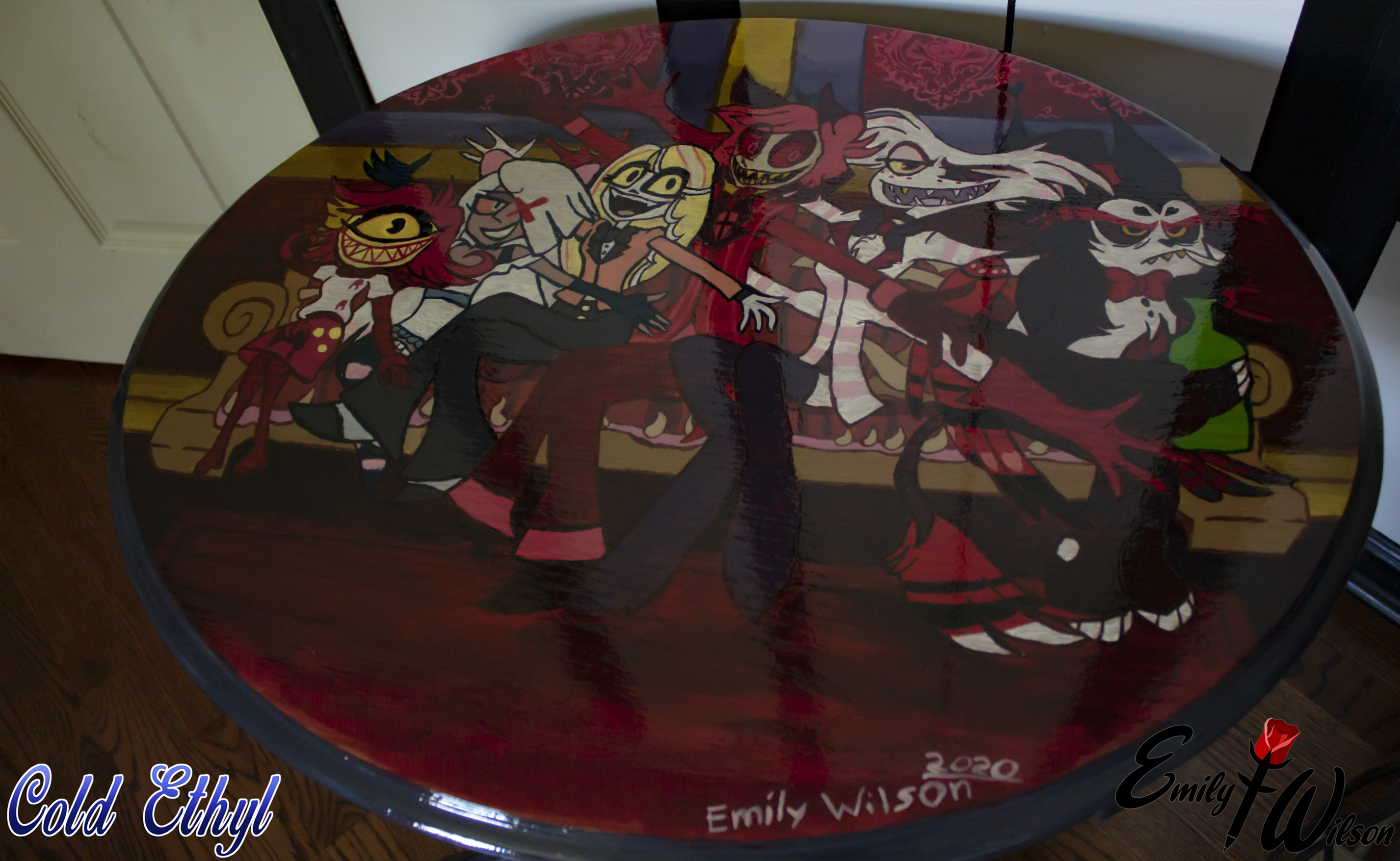 Photogaph depicting a table surface with Hazbin Hotel fanart. The characters are sitting on a couch together with their arms and hands sticking out. Left to right: Niffty (whose doing the Baphomet hand pose), Vaggie, Charlie, Alastor, Angel, and Husk. (Whose turned away and not doing the hand pose. He's holding a green bottle of booze looking annoyed.)